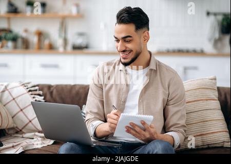 Positive smart indian or arabian man taking notes while working on project or learning online uses laptop, sitting on a sofa in a living room, browsing information in internet, prepares a report Stock Photo