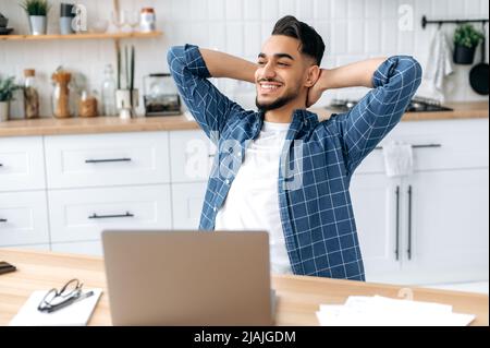 Happy arabian or indian guy, freelancer, working from home, sits in the kitchen at the workplace, takes a break from work, puts his hands behind his head, looks to the side, dreams of rest, smiles Stock Photo