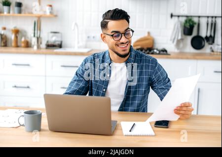 Smart successful arabian or indian guy with glasses, freelancer or office worker working remotely, sit at a desk at home in the kitchen, working on a project uses laptop, studying documents, smiling Stock Photo