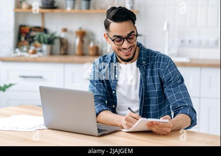 Happy charismatic arabian or indian guy with glasses, sitting at the desk at home, taking notes in a notebook during an online webinar or lecture, using a laptop, smiling, gaining knowledge Stock Photo