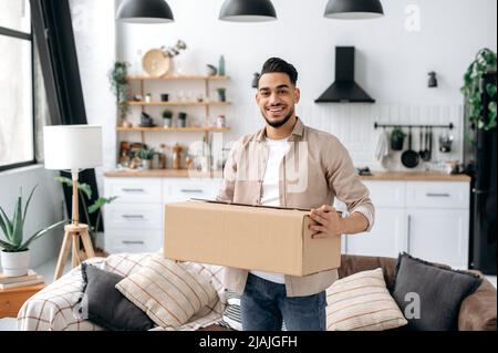 Joyful stylish indian or arabian guy, stand at home in the living room, holding a large cardboard box, received a long-awaited parcel from the online store, preparing to unpack, looks at camera,smiles Stock Photo