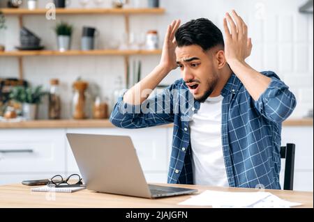Angry stressed arabian or indian guy, sitting at desk in the kitchen, using laptop for online work, annoyed with bad result of his project, failed the deal, experiences negative emotions and stress Stock Photo