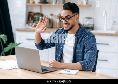 Positive indian or arabian guy, online tutor, freelancer, student, wearing glasses and casual clothes, speaking via video conference, looking at laptop, conducts virtual meeting, work from home,smile Stock Photo