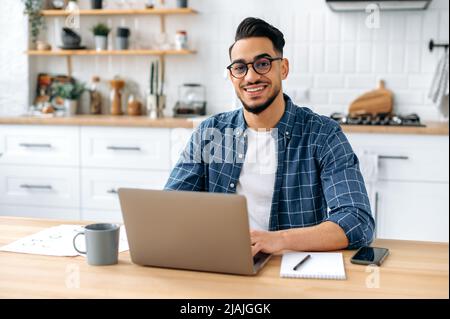 Portrait of a handsome successful confident Arabian or Indian guy with glasses, freelancer, designer or IT specialist, sitting at home in the kitchen, working online by laptop, looks at camera, smiles Stock Photo