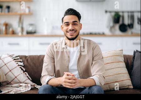 Portrait of an attractive handsome indian or arabian guy in a stylish casual wear, sitting on sofa at home in a living room, in a stylish interior, looking at the camera, smiling positively Stock Photo