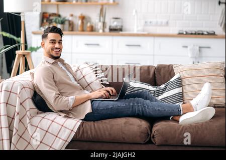 Attractive smiling arabian or indian man in casual stylish wear, freelancer or student, sitting on couch in living room, working on a project uses laptop, looking at the camera, smiling happily Stock Photo