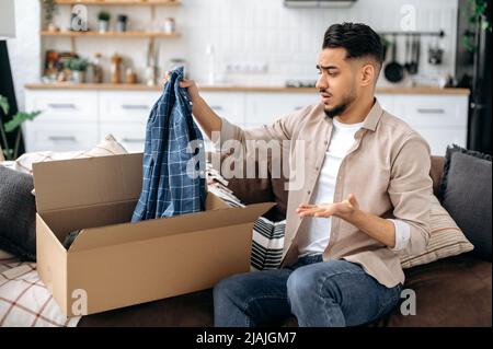 Sad unhappy indian or arabian guy opening big carton box, unpacking internet store order at home, feels frustrated with long delivery service and the goods which received, experiencing confusion Stock Photo