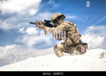 Man in military camouflage uniform with service rifle replica, standing on top of sand dune with cloudy sky in background. Stock Photo