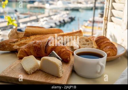 French breakfast with fresh baked croissants, baquett bread, crottin goat cheese, black coffee and view on fisherman's boats in harbour of Cassis, Pro Stock Photo