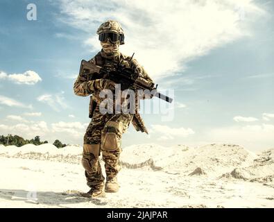 Serviceman in camouflage uniform running with Army service rifle in desert area. Stock Photo