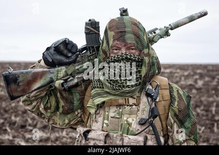 Army sniper standing in field with rifle, face hidden behind shemagh. Stock Photo