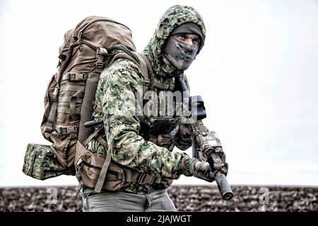 Military mercenary wearing hooded camo jacket and backpack, walking through muddy terrain. Stock Photo