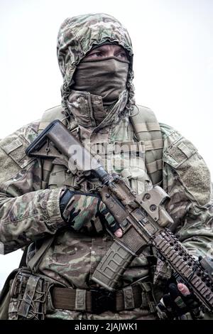 Military mercenary wearing hooded camo jacket and backpack, armed with rifle, looking away. Stock Photo