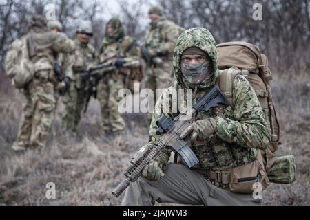 Infantry soldier wearing hooded camo jacket and backpack, kneeling with firearm in hands. Stock Photo