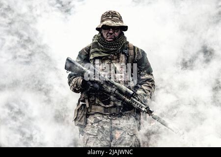 Elite commando fighter in camouflage uniform, armed with sniper rifle, standing in clouds of smoke. Stock Photo