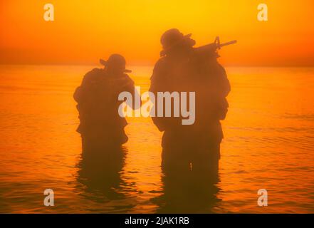 Silhouette of two soldiers wading the shoreline, sneaking in the darkness with assault rifles drawn. Stock Photo
