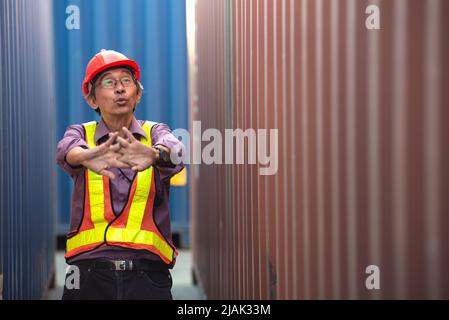 Senior elderly asian worker engineer wearing safety vest and helmet, raising hand up, standing between containers at logistic shipping cargo container Stock Photo