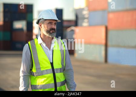 Caucasian male manager in white hard hat helmet and high-visibility vest walking in Container Terminal. Stock Photo