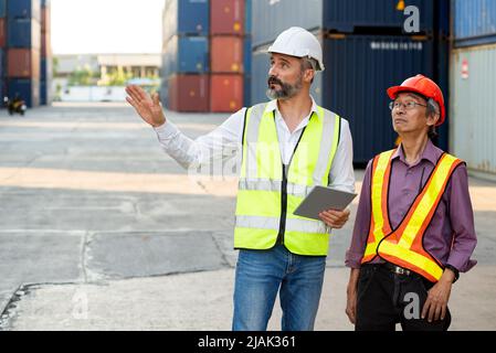 Caucasian male manager and senior engineer use tablet to check import export cargo containters loading at storage distribution warehouse. Stock Photo