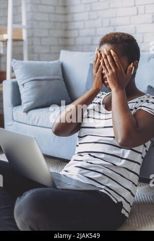 Stressed afro woman grabbing head and using laptop, while sitting on the floor. Beautiful african woman. Woman working on laptop. Stock Photo
