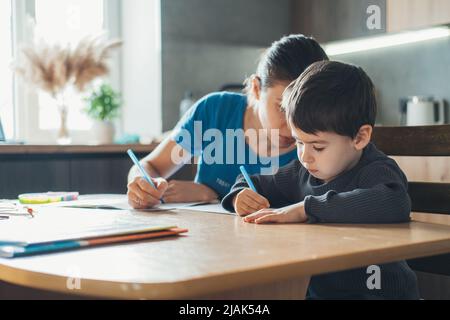 Mother and son writing in notepad while sitting together at table and helping son to do homework. Homework assignment. Adult woman teaches the child Stock Photo