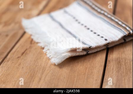 A white kitchen towel lies on the countertop of a wooden table. The towel is fringed and patterned on the rough boards. Selective Focus. Stock Photo