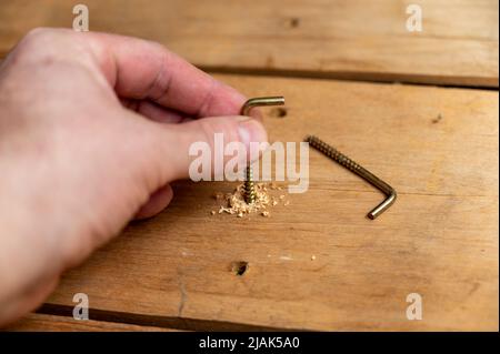 A man's hand screws a screw into a board. Screw with an L-hook. Galvanized screw. Drilled hole in the board. Selective focus. Stock Photo