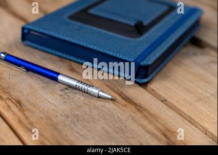A blue diary and pencil against a wooden background. A closed hardcover notebook. A metallic silver-blue pencil rests on top of old cracked boards. Cl Stock Photo
