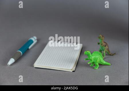 Two Dinosaur miniatures, notebook and ballpoint pen against a gray background. Small green figures of animals of prey standing on their hind legs. Spr Stock Photo