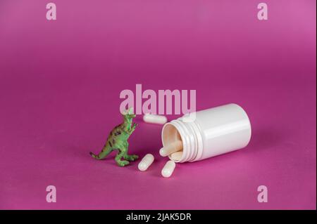 A white vial with pills spilling out and a miniature dinosaur against a purple background. Small green figure of a predatory animal standing on its hi Stock Photo