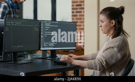 Portrait of focused programer writing code looking at multiple computer screens displaying programming language algorithm. Software developer coding while colleagues doing teamwork in background. Stock Photo
