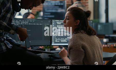Junior programer sitting down on desk showing laptop with source code to senior dev asking for opinion. Software developer writing algorithm interrupted by colleague wanting help with fixing errors. Stock Photo