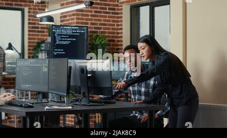 Two software developers holding laptop with coding interface walking towards desk and sitting down talking about group project. Programmers team discussing algorithms pointing at computer screen. Stock Photo