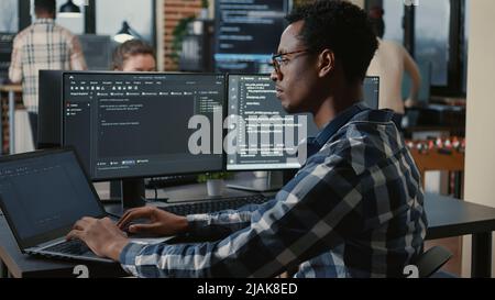 Focused programer fixing glasses while typing on laptop sitting at desk with multiple screens parsing code. Coder working on user interface with team of programmers coding big data in the background. Stock Photo