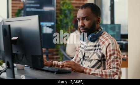 Portrait of focused african american programer wearing wireless headphones working looking at computer screen while typing. Software developer writing code in it agency office. Stock Photo