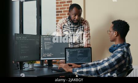Software developer sitting at desk with multiple screens and laptop running code talking with colleague about artificial intelligence algorithm. Software developers doing teamwork in it office. Stock Photo