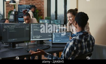Software developers celebrating successful code compiling doing high five hand gesture with colleague. Programmer analyzing algorithm on multiple screens takes off glasses and congratulates coworker. Stock Photo
