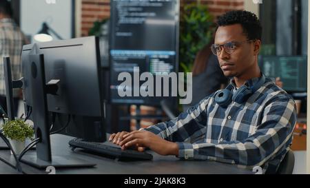 Portrait of focused programer writing code fixing glasses and smiling sitting at desk in it startup. Coder programming user interface with team of programmers working in the background. Stock Photo