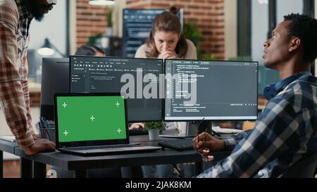 Programer taking glasses off talking with colleague at desk with laptop with green screen chroma key mockup and multiple computer screens compiling running code. Coders doing online cloud computing. Stock Photo