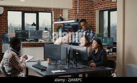 Software engineers brainstorming ideas looking at code on computer screens asking feedback from senior developer while intern joins the discussion. Junior programmers collaborating on group project. Stock Photo