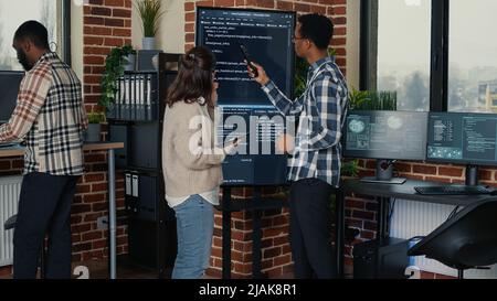 Team of database admins analyzing source code on wall screen tv comparing errors using digital tablet in busy server room. Two cloud programers debugging algorithm in software innovation office. Stock Photo