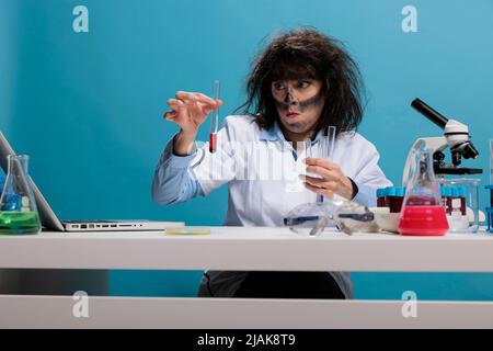 Foolish crazy looking mad chemist sitting at desk in laboratory while experiment with new chemical compounds after lab explosion. Maniac biochemistry specialist with dirty face testing substances. Stock Photo