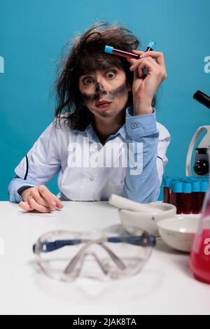 Potrait of crazy looking mad silly chemist with messy hair and dirty face having glass test tubes filled with blood samples after laboratory explosion. Maniac scientist sitting at desk. Studio shot Stock Photo