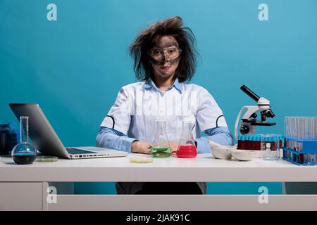 Silly looking crazy chemist with messy hair and dirty face sitting at desk after failed laboratory experiment explosion. Foolish lab worker with wild appearance sitting on blue background. Studio shot Stock Photo