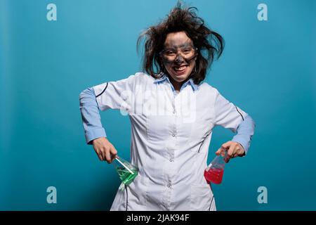 Lunatic biochemistry specialist grinning creepy while holding Erlenmeyer flasks filled with liquid substances. Insane looking mad chemist with dirty face and messy hair acting goofy on blue background Stock Photo