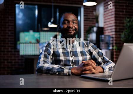 Forex stock market analyst in trading agency workspace sitting at table while smiling at camera. Investment agency confident and professional employee sitting at desk in office. Stock Photo