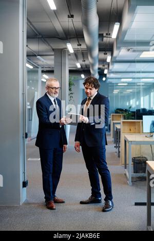 Two businessmen in formal wear discussing online presentation on digital tablet while standing at office Stock Photo