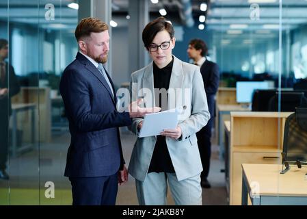Young businessman pointing at business contract and discussing it together with businesswoman while they standing at office Stock Photo