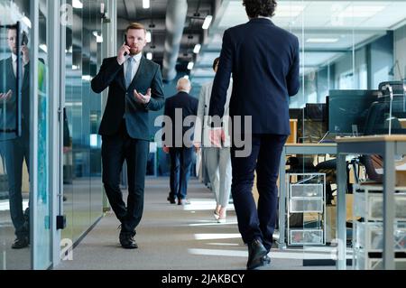 Young businessman in suit talking on mobile phone while walking along office corridor among business people Stock Photo