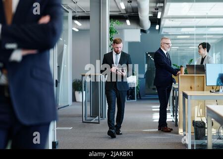 Young businessman using tablet pc walking along corridor with business people in formal clothes working in office Stock Photo